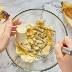 Ręce ugniatają banany w misce, przygotowując pyszny deser z bananów. Obok leżą dojrzałe banany.