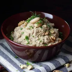 Aksamitna pasta z lubczyku, udekorowana fasolką, orzechami i ziołami, w bordowej misce na pasiastym obrusie.
