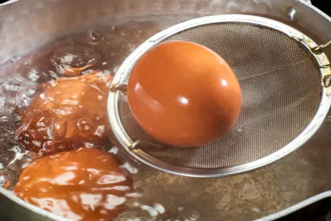 Jajka na miękko czas! Brązowe jajka gotują się w bulgoczącej wodzie, jedno z nich jest wyciągane sitkiem.
